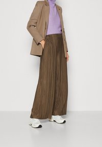 Bruine plooien wijde broek gecombineerd met een aansluitende paarse coltrui en een beige blazer. Model draagt witte en zwarte sneakers.