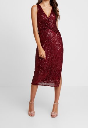 Kobieta w bezrękawnikowej, głęboko czerwonej sukience midi z cekinami, z dekoltem w serek i beżowych sandałkach na wysokim obcasie z paskami, stoi na jednolitym tle.