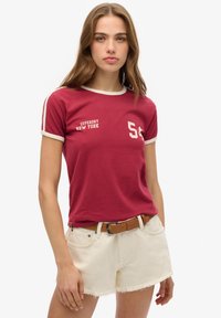 Rød t-shirt med hvid kant, der har teksten "SUPERDRY NEW YORK" og nummeret '56.' Sammen med hvide denim shorts med slidte kanter.