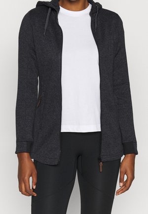 Person iført en mørkegrå lynlåshoodie over en hvid t-shirt og sorte leggings, stående med armene afslappet ved siden.