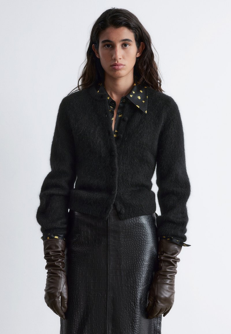 Schwarze Strickjacke mit strukturiertem Finish, Knopfverschluss und langen Ärmeln über einem gemusterten schwarzen Hemd. Ein Paar braune Lederhandschuhe getragen.