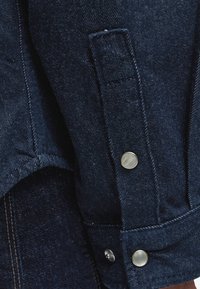 Κοντινή εικόνα σκούρου μπλε τζιν ύφασμα με μεταλλικά κουμπιά και λεπτομερείς ραφές σε ενδύματα.