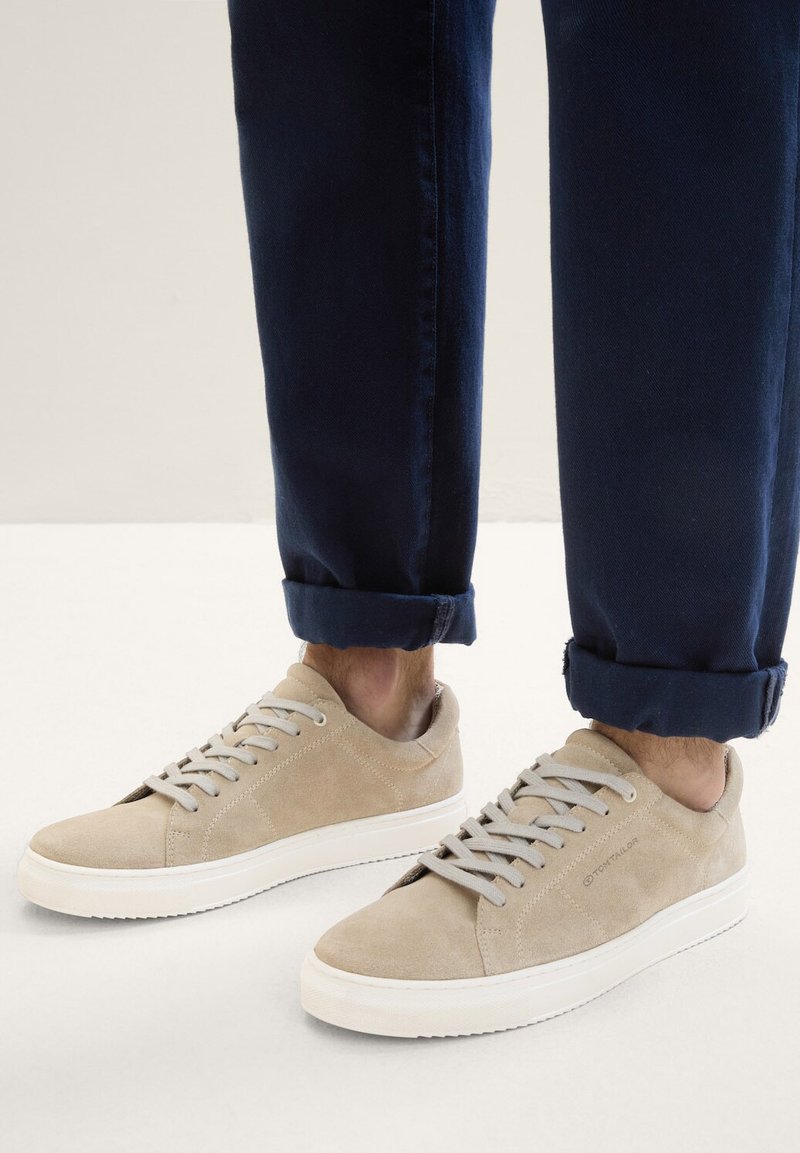 Zapatillas de ante beige con cordones grises y suela de goma blanca. Pantalones azul marino enrollados que revelan los zapatos; textura suave y diseño casual.