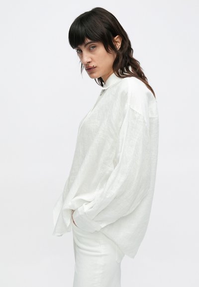 Λευκό πουκάμισο από λινό σε μεγάλο μέγεθος με όρθιο γιακά, φαρδιά μανίκια και υφή στο ύφασμα. Συνδυάζεται με λευκό παντελόνι για ένα αρμονικό σύνολο.