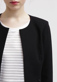 Veste noire courte avec un col rond et sans boutons visibles, portée sur une chemise blanche à rayures avec un tissu texturé.