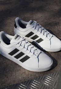 Baskets blanches Adidas avec des bandes et des lacets noirs, placées côte à côte sur une surface en béton avec une grille métallique à proximité.