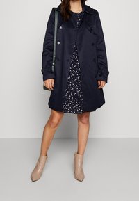 Marinblå trenchcoat med dubbelknäppning, bärs över en marinblå klänning med vita blommönster. Beige ankelboots med spetsiga tår.