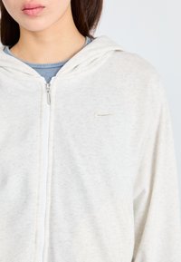 Svijetlosivi Nike dukserica sa prednjim zatvaračem, mekane teksture, s izvezbom logotipa na lijevoj strani grudi. Ima kapuljaču i standardni kroj.