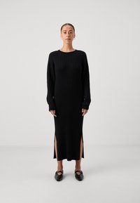 Črna pletena obleka z dolgimi rokavi, stranskimi razporki na robu, okroglim izrezom in gladko teksturo. Ujemajoči se črni ravni čevlji.