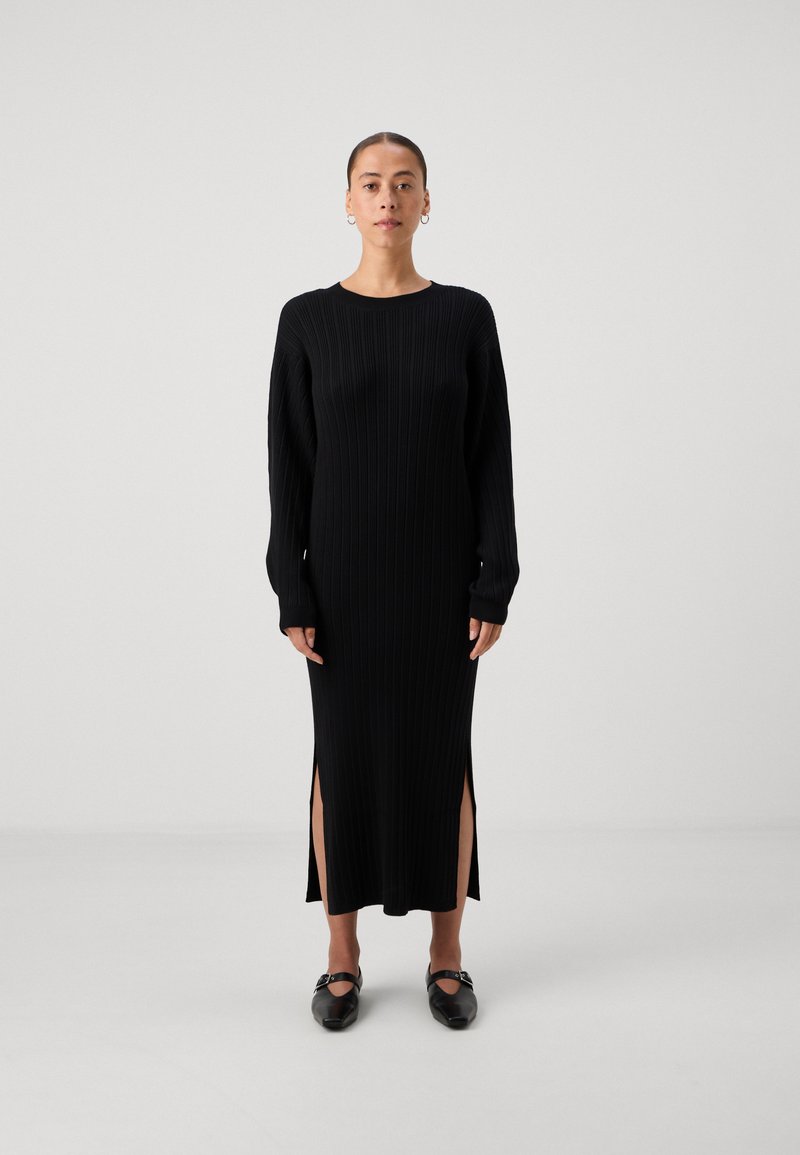 Črna pletena obleka z dolgimi rokavi, stranskimi razporki na robu, okroglim izrezom in gladko teksturo. Ujemajoči se črni ravni čevlji.