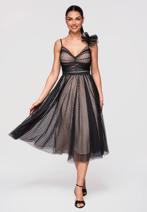 Kobieta nosząca czarną, przezroczystą, midi sukienkę bez rękawów w groszki, z kwiatowym detalem na ramieniu, uśmiechająca się i trzymająca rozłożoną spódnicę.