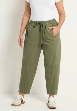 Femme portant un pantalon à cordon de couleur vert olive, un haut blanc à manches courtes, et des baskets blanches avec des rayures noires, debout devant un fond neutre.