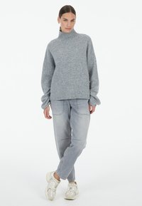 Grauer gestrickter Pullover mit hohem Kragen, lockerer Passform und Perlenakzenten. Kombiniert mit hellgrauen weiten Jeans und grauen Sneakers. Flachter Hintergrund.