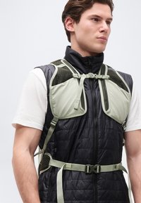 Noor mees, kes kannab valge särgi üle musta tikandiga vestit, seljas helerohelise rakmestiilis matkakoti rihm, mis on lukustatud rinna ja vöökoha kohal.