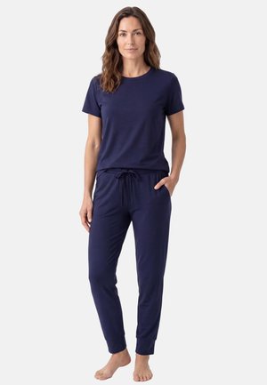 Vrouw staat op blote voeten, draagt een donkerblauw T-shirt met korte mouwen en bijpassende joggingbroek met trekkoord, tegen een effen lichte achtergrond.