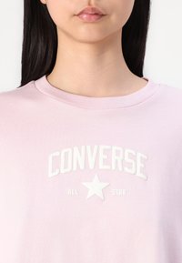 Lys rosa bomull t-skjorte med hvit preget "CONVERSE ALL STAR" tekst og stjernedesign. Rund hals og glatt tekstur.