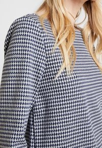 Marinblå och vit mönstrad tyg, med ett houndstooth-mönster och en texturerad yta. Plagget har en lös passform och en avslappnad halsringning.