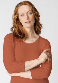 Maglietta a maniche lunghe color ruggine in un tessuto morbido e liscio con scollo rotondo. Il modello ha le braccia incrociate, mostrando un fit rilassato.