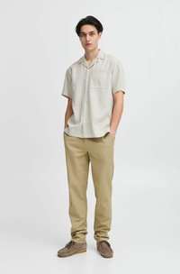 Kurze, cremefarbene Hemd mit einem subtilen gepunkteten Muster, kombiniert mit beigen Hosen und braunen Wildlederschuhen. Der Look ist entspannt und lässig.
