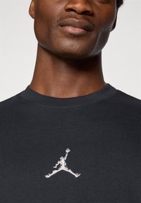Svart bomull T-skjorte med rund hals og brodert hvit Jumpman-logo på venstre bryst. Glatt tekstur, middels passform.