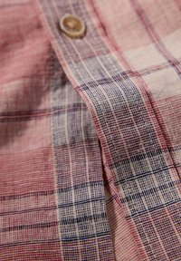 Geweven shirtstof met een geruit patroon in rood, marineblauw en beige, met een zichtbare knoopdetail en een gestructureerde weving.