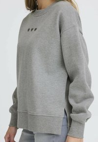 Sudadera gris con mangas largas, cuello redondo, aberturas laterales y pequeño estampado de corazón en el pecho. Fabricada con un tejido suave de textura lisa.