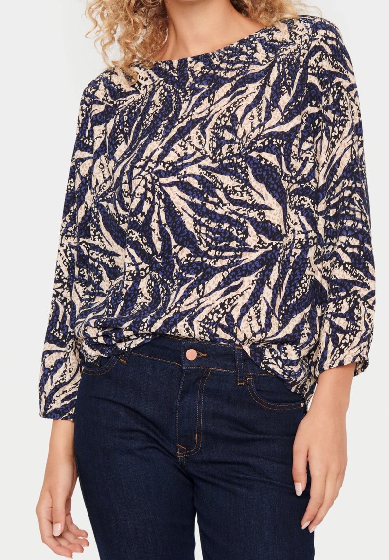 Top w kwiatowy wzór w granatowym i kremowym kolorze, z długimi rękawami i luźnym krojem, wykonany z lekkiego materiału; zestawiony z ciemnymi dżinsami.