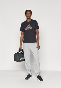 Črna majica s kratkimi rokavi z leopardjim printom Adidas logotip, sivi trenirka, črne športne čevlje, drži črno potovalno torbo z belim Adidas logotipom.