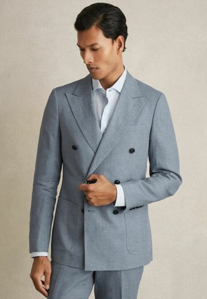 Uomo che sistema un bottone sulla giacca doppiopetto grigio chiaro indossata sopra una camicia bianca, in piedi davanti a uno sfondo beige uniforme.