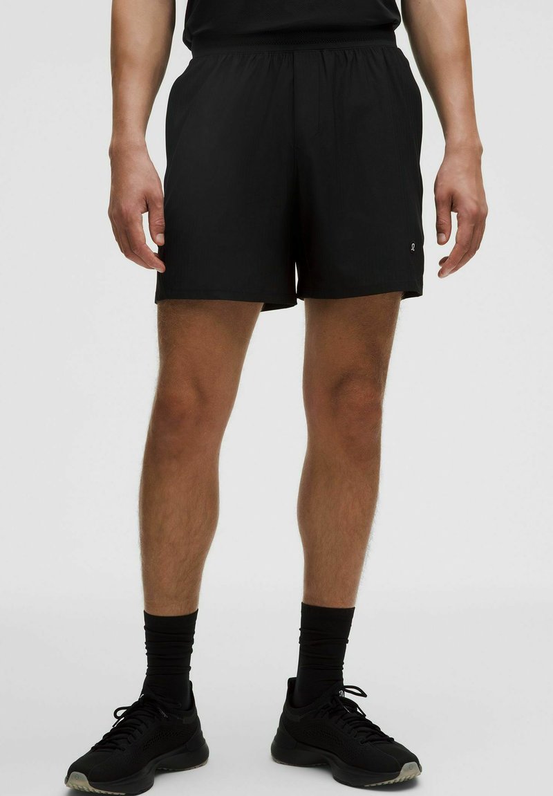 Pantalones cortos deportivos negros hechos de tela ligera, con cinturilla elástica y un detalle sutil de logo. La longitud llega a la mitad del muslo.