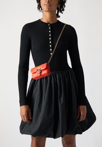 Zwarte geribde jurk met lange mouwen en een uitlopende rok, gecombineerd met een kleine rode leren handtas met een gevlochten schouderband en sluiting.