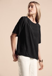 Maglietta nera a maniche corte con scollatura rotonda e dettagli in filo bianco con un motivo ondulato. Indossata con pantaloni bianchi, che presentano una vestibilità comoda.