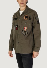 Olijfgroene militaire stijl jas met meerdere geborduurde patches, knoopsluiting en borstzakken. Katoenen stof met een gladde textuur.