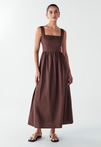 Rjava maksi obleka s kvadratnim izrezom, širokimi naramnicami in nabranim pasom. Oblečena z črnimi sandalmi z zlatimi detajli. Gladka tekstura.