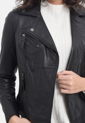 Frau trägt eine schwarze Lederjacke über einem weißen Strickpullover und hält den Reißverschluss der Jacke mit der rechten Hand.