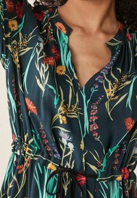 Robe fleurie en tissu vert foncé avec un décolleté en V, des manches courtes bouffantes et une taille cordon. Le motif comprend des fleurs et des feuilles multicolores.