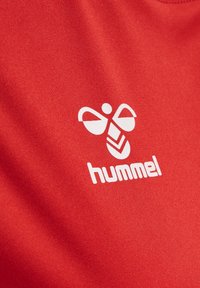 Logo d'abeille stylisé blanc et texte "hummel" imprimés sur un tissu rouge texturé.