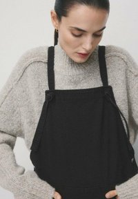 Schwarzer Jeans-Overall mit verstellbaren Trägern, getragen über einem hellgrauen, grob gestrickten Pullover. Der Stoff zeigt Textur und eine entspannte Passform.