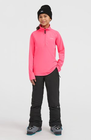 Roze fleece top met kwartrits, zwarte geïsoleerde sneeuwbroek en grijze sneeuwlaarzen met blauwe accenten. Het model draagt een zwarte beanie.
