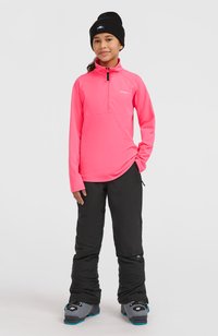 Roze fleece top met kwartrits, zwarte geïsoleerde sneeuwbroek en grijze sneeuwlaarzen met blauwe accenten. Het model draagt een zwarte beanie.