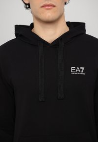 Čierna bavlnená mikina s kapucňou a šnúrkami, na hrudi s bielym logom EA7. Tkanina vyzerá hladko a pohodlne.