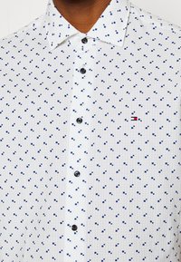 Camicia bianca con bottoni, caratterizzata da un piccolo motivo a quadri blu ripetuto. Bottoni neri e un accento rosso e blu nel logo sul petto.