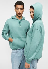 Δύο άτομα που φορούν ταιριαστά φούτερ με κουκούλα σε χρώμα μέντας, κατασκευασμένα από απαλό ύφασμα. Τα φούτερ διαθέτουν μπροστινή τσέπη και κορδόνι προσαρμογής.