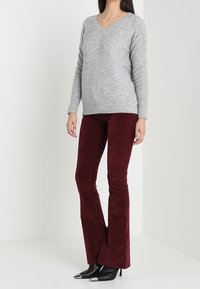 Un pull en tricot gris avec un col en V, associé à un pantalon en velours côtelé bordeaux évasé. Le modèle porte des bottines à talons noires.