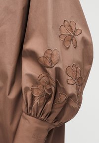 Καφέ υφασμάτινο μανίκι με μανσέτα μαζεμένη, με ραμμένα μοτίβα κοπής λουλουδιών και διακριτική σήμανση MAX&Co. κοντά στη μανσέτα.