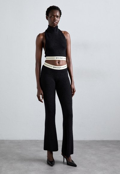 Tall woman wearing black sleeveless crop top and matching pants with cream elastic bands labeled "Palm Angels," standing on gray floor.
