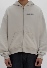 Ljusgrå huvtröja med dragkedja, utrustad med en framficka, ribbade muddar och en huva med en enkel, broderad logotyp.