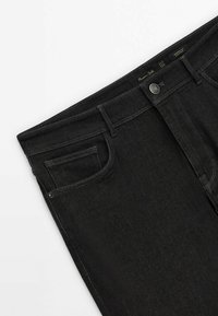 Schwarze Jeans mit schmal zulaufender Passform, vorderer Münztasche, Gürtelschlaufen, Metallknopf und sichtbarem Markenlabel innen im Bund.
