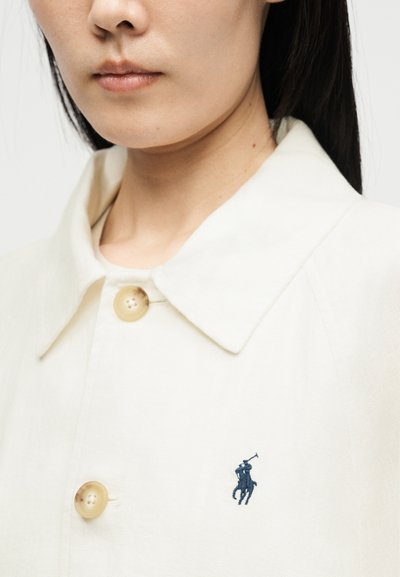 Záber zblízka osoby, ktorá má na sebe bielu košeľu s golierom, béžovými gombíkmi a malým tmavomodrým vyšívaným logom hráča póla.