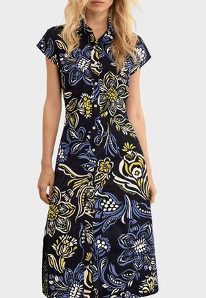 Donna che indossa un vestito nero a maniche corte con bottoni e grandi motivi floreali blu, bianchi e gialli.